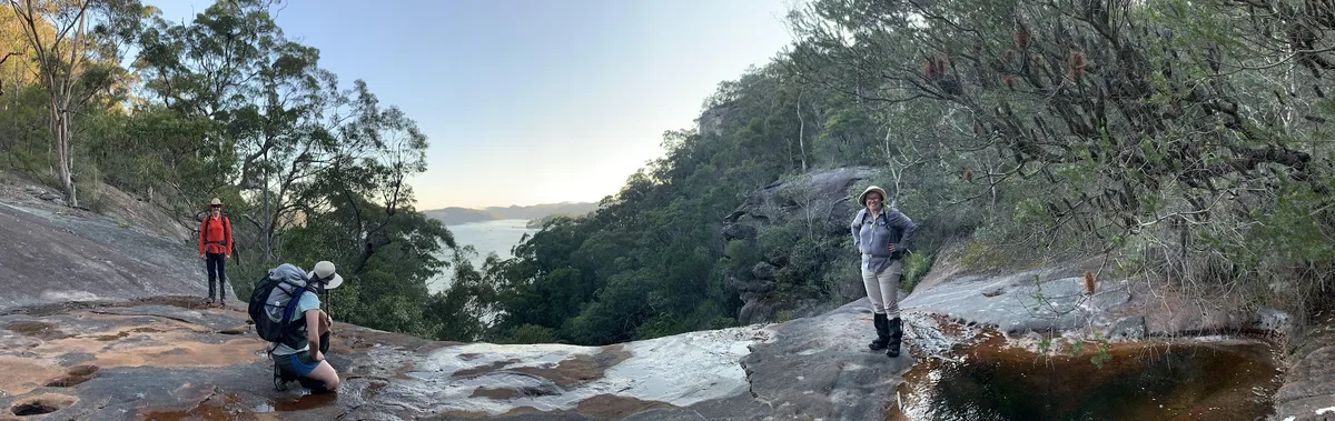 Three hikers on a sandstone ledge overlooking the Hawkesbury River at sunrise