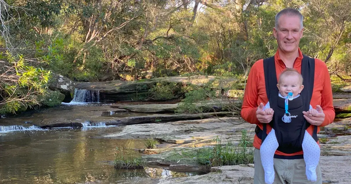 Father carrying a smiling baby beside a cascading waterfall on the Piles Creek loop