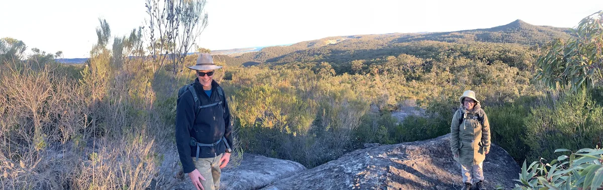 Two hikers pause on a rock ledge with Mount Wondabyne's peak in the distance