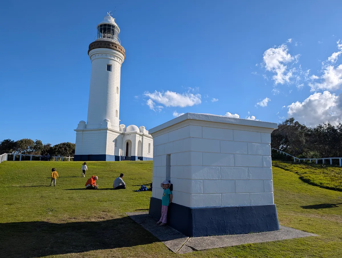 Families relaxing on the lawn below Norah Head Lighthouse with a small heritage signal hut in the foreground