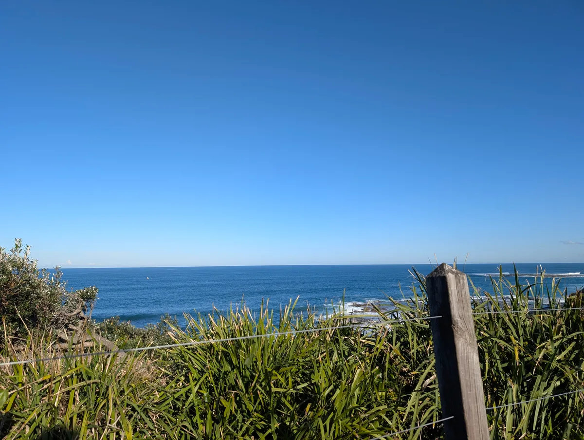 The ocean and rock shelf seen from the nature trail at Norah Head, with coastal grasses and a weathered fence post in the foreground