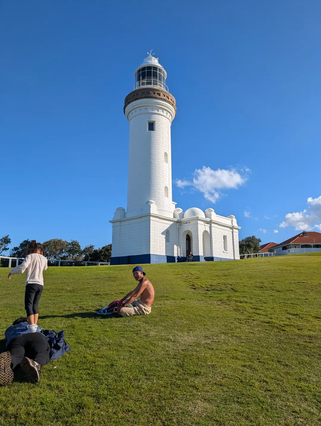 Norah Head Lighthouse with visitors resting on the grassy slope in the afternoon light