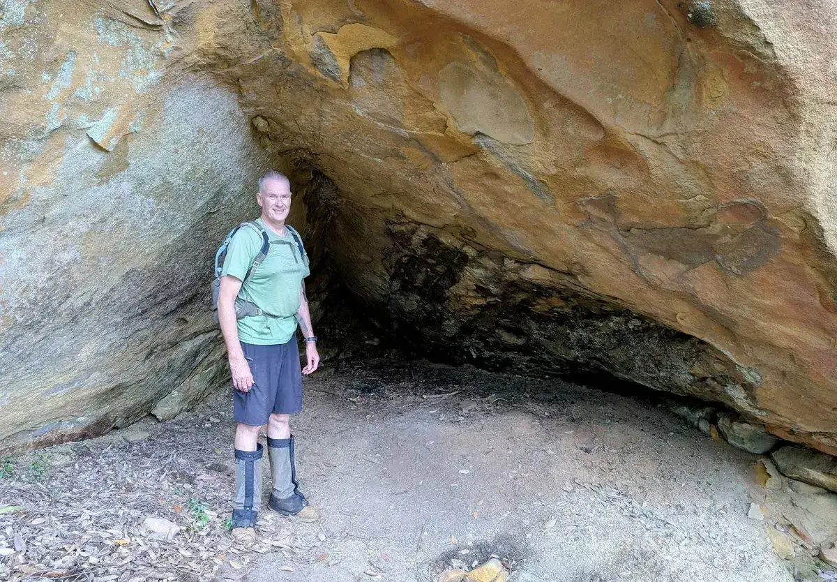 Hiker smiles at the mouth of a sandstone cave wearing gaiters