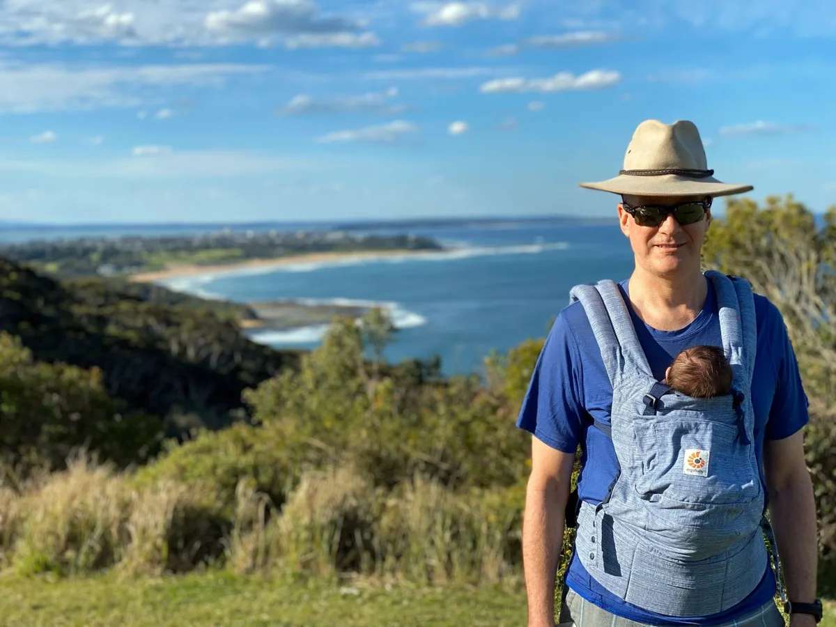 Father carrying a baby in a carrier beside a gnarled coastal tree